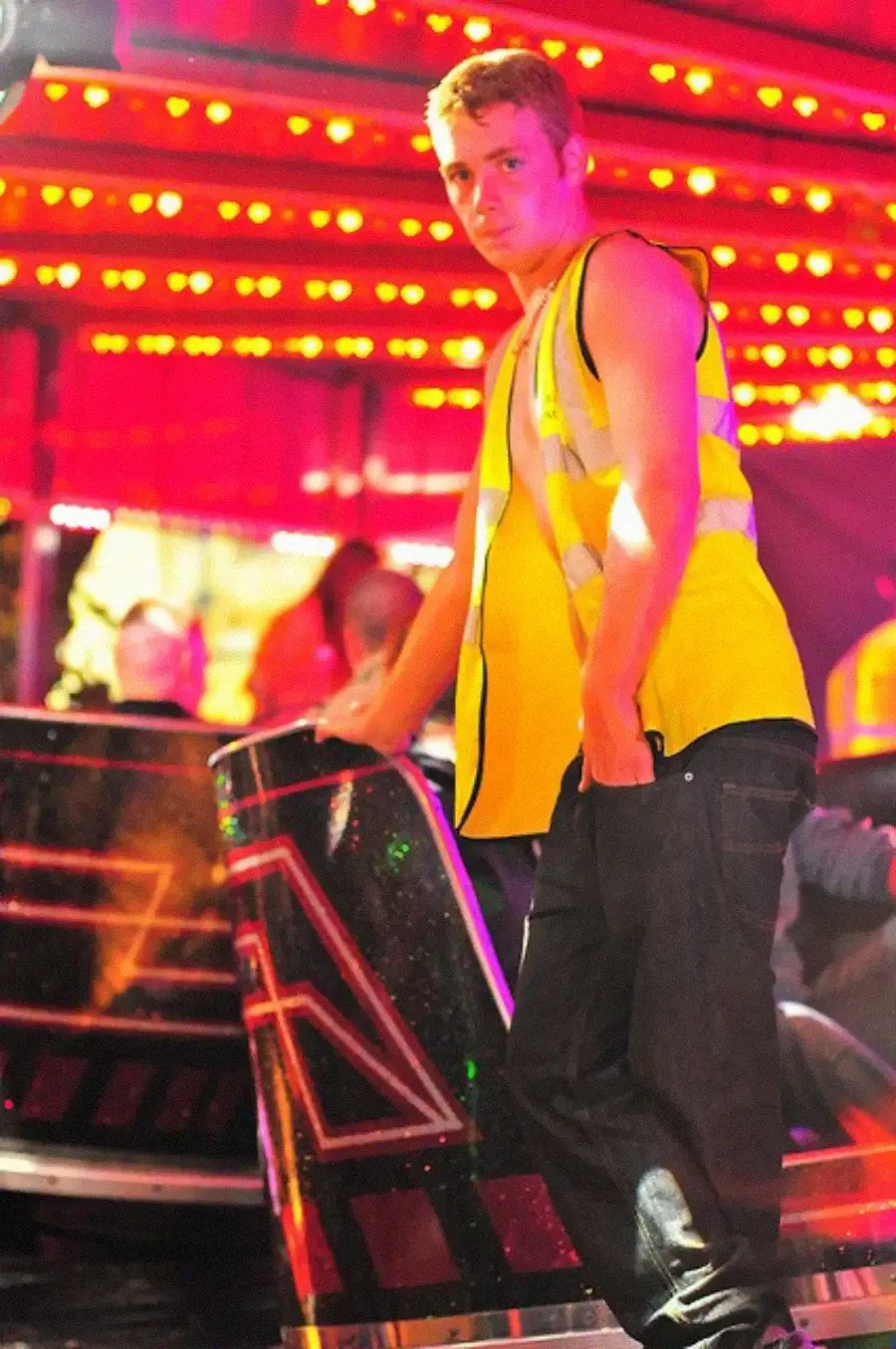 A young man in a yellow reflective vest stands beside a fairground ride under bright red and yellow lights, looking at the camera with one hand in his pocket.