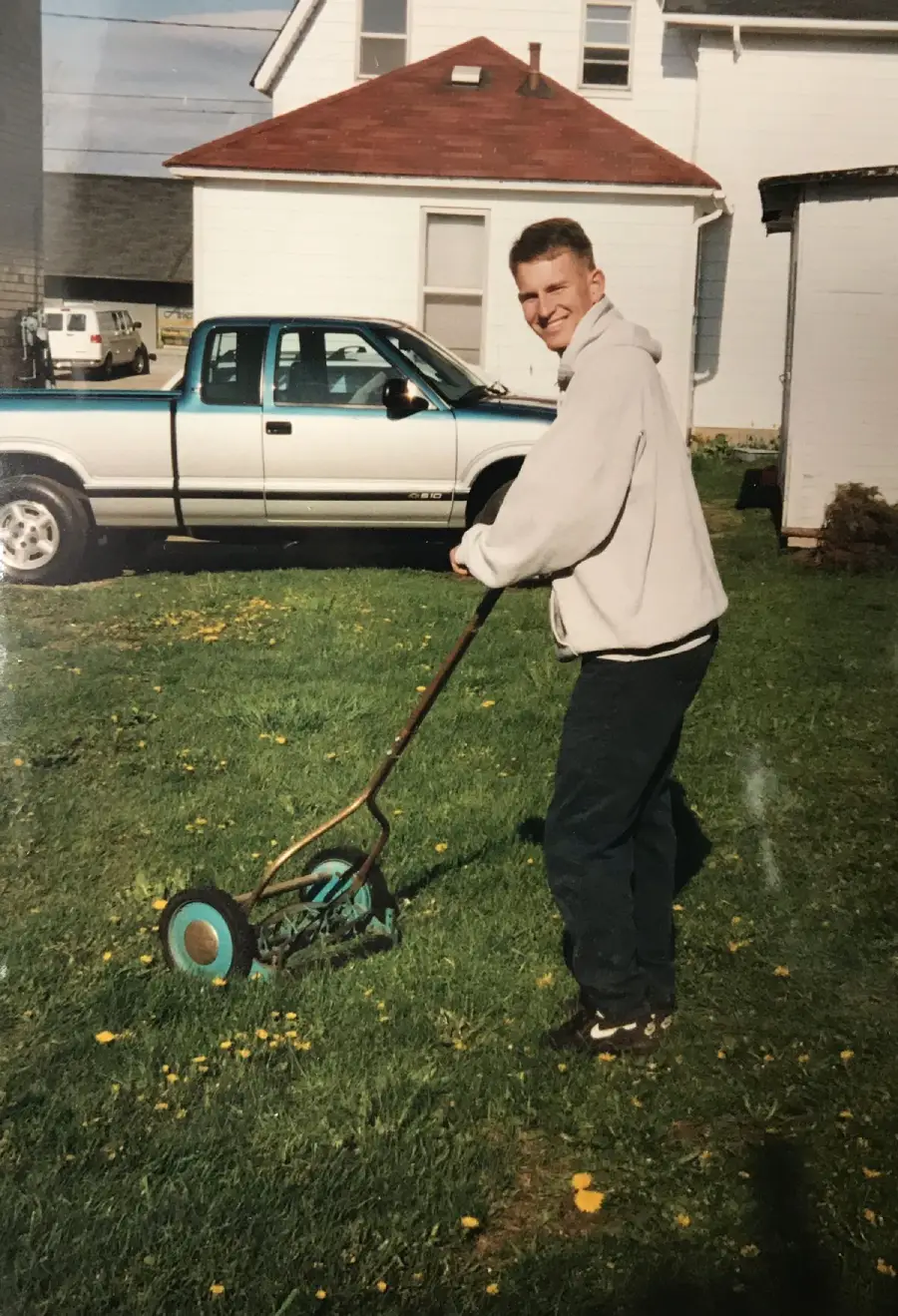 A young man wearing a gray hoodie and dark pants is mowing the lawn with a manual push mower in a yard. There is a white pickup truck and a house in the background.