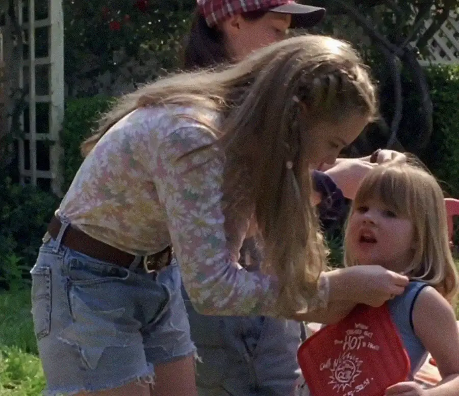 A woman in a floral top and denim shorts helps a young blonde girl adjust a red bib outdoors, while another person wearing a plaid cap stands in the background.