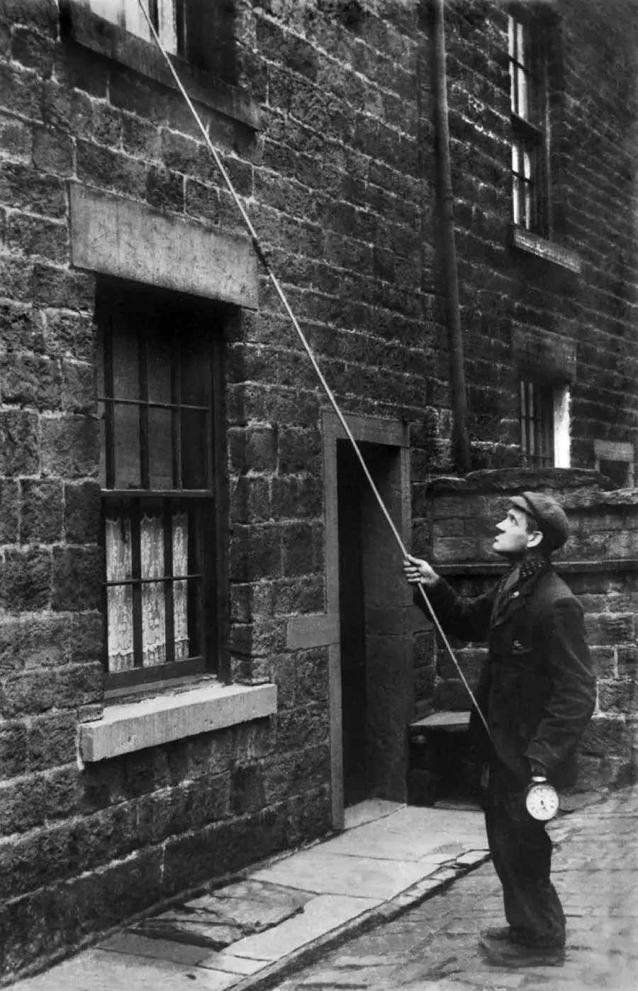 A man on a cobblestone street uses a long pole to tap on an upstairs window of a brick building, possibly to wake someone. He wears a cap and holds a timepiece, suggesting his role as a knocker-upper.