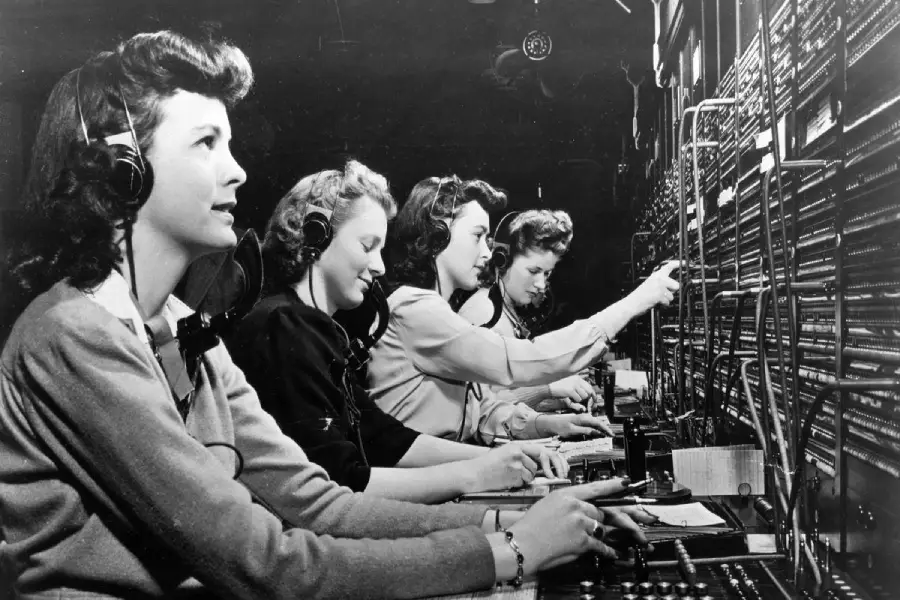 Four women in vintage attire sit in a row at a large telephone switchboard, wearing headsets and connecting calls by plugging in cables. The scene is black and white, suggesting an era from the mid-20th century.