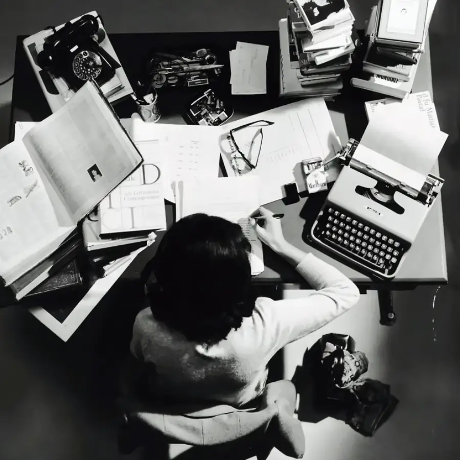 A person sits at a cluttered desk covered with papers, books, a typewriter, a rotary phone, and office supplies, seen from above. They appear to be writing or working intently.