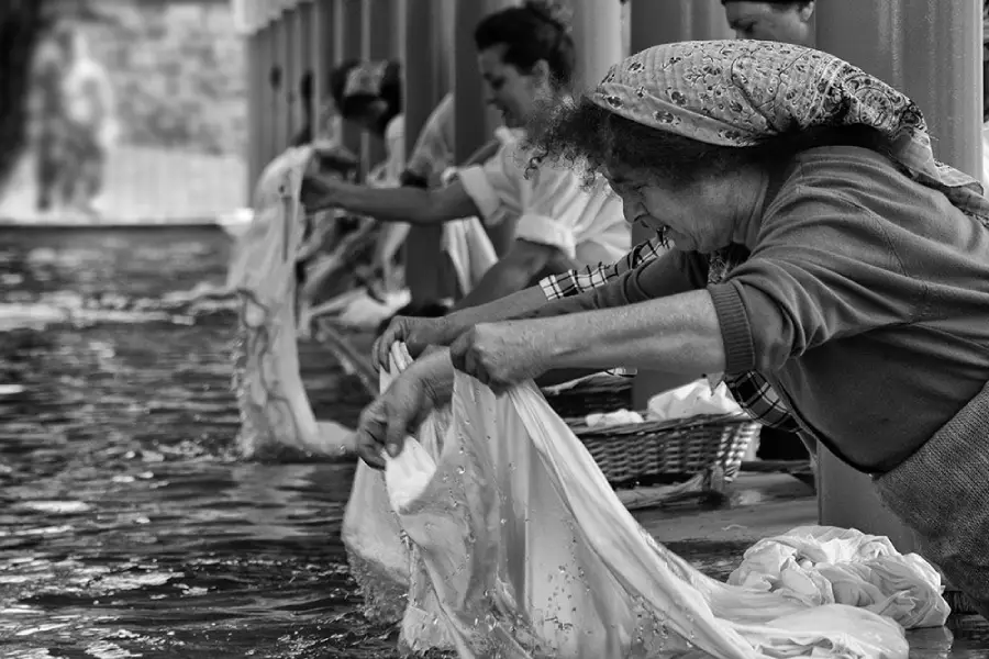 Women wash clothes by hand in a river or washhouse, wringing out wet fabric. The image is black and white, showing focus and effort on their faces as they work together in a line.
