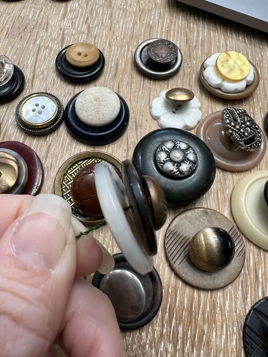 A hand holds a stack of layered vintage buttons on a wooden surface, with more assorted, decorative buttons spread out in the background.