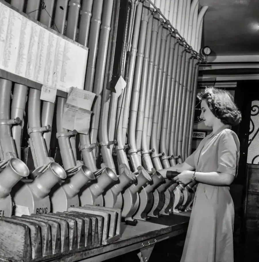 A woman in a dress stands by a wall of large tubes labeled with initials, sorting mail into the tubes in what appears to be an old postal sorting facility. Papers and a chart hang above the tubes.