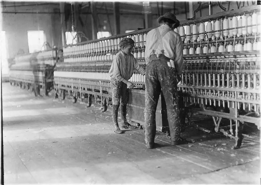 A black-and-white photo shows a boy and a man working at a long row of spinning machines in a textile mill, with spools of thread visible. The factory setting appears old, with wooden floors and industrial equipment.