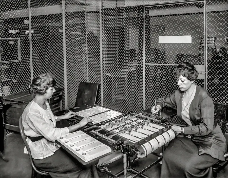 Two women sit at a table operating a vintage machine with paper rolls and control levers in a fenced industrial workspace, possibly performing data processing or calculating tasks.