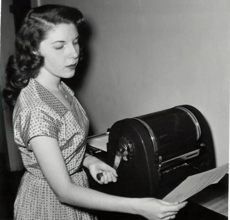A woman in a patterned dress stands beside a vintage mimeograph machine, holding a freshly printed sheet of paper and looking at it thoughtfully.