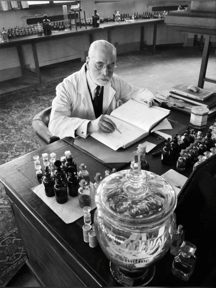 An older man in a lab coat writes in a large book at a desk filled with glass bottles and scientific equipment in a laboratory setting.