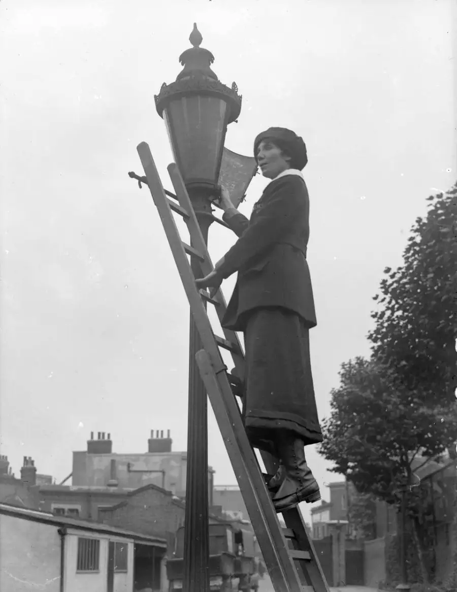 A woman wearing a long coat and hat stands on a ladder, lighting or cleaning a streetlamp in an urban area with buildings and trees in the background. The image is black and white.
