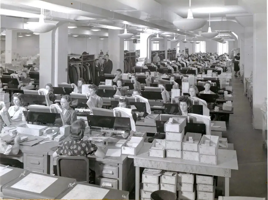 Black-and-white photo of a large, crowded office filled with rows of desks where men and women work with papers and boxes. Coat racks and shelves are visible along the walls. The atmosphere appears busy and organized.