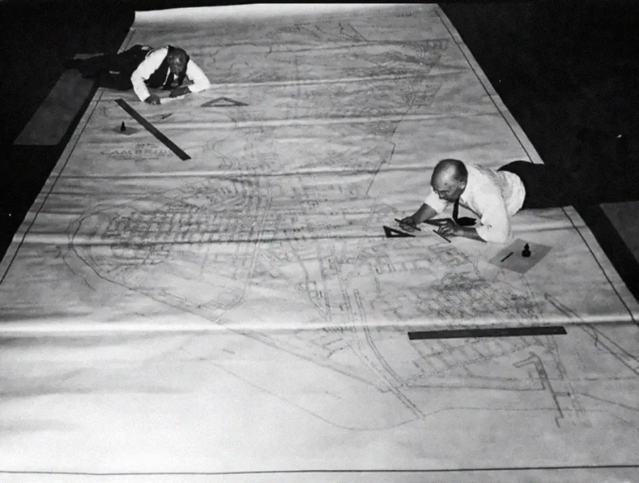 Two men in dress shirts and ties lie on a giant blueprint, using rulers and drawing tools to work on the large architectural plan spread out on the floor.