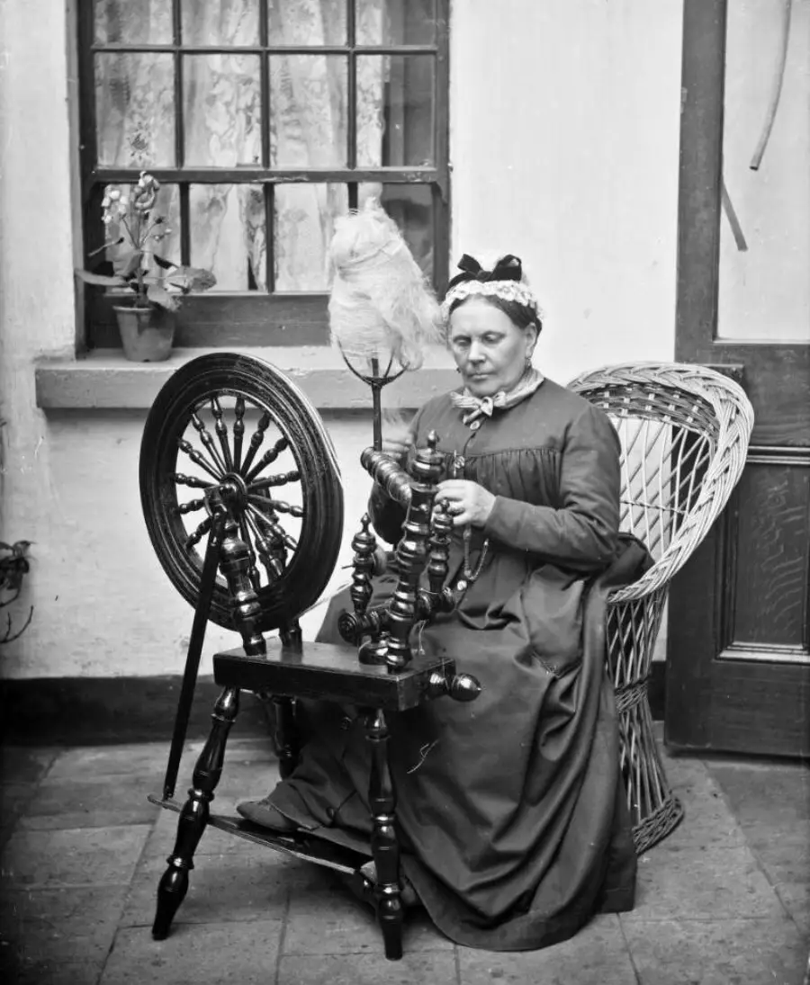 A woman in a long dress and headscarf sits in a wicker chair, spinning wool on a spinning wheel indoors near a window with potted plants.
