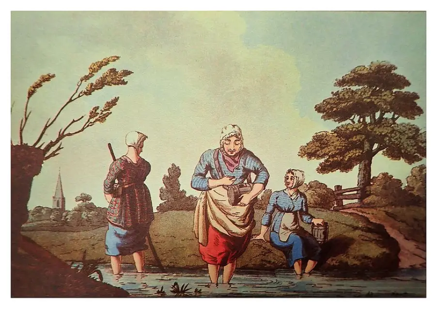 Three women in old-fashioned clothing stand in a shallow stream, washing clothes. Trees and a fence are in the background, with a church steeple visible in the distance on the left. The scene appears pastoral and calm.