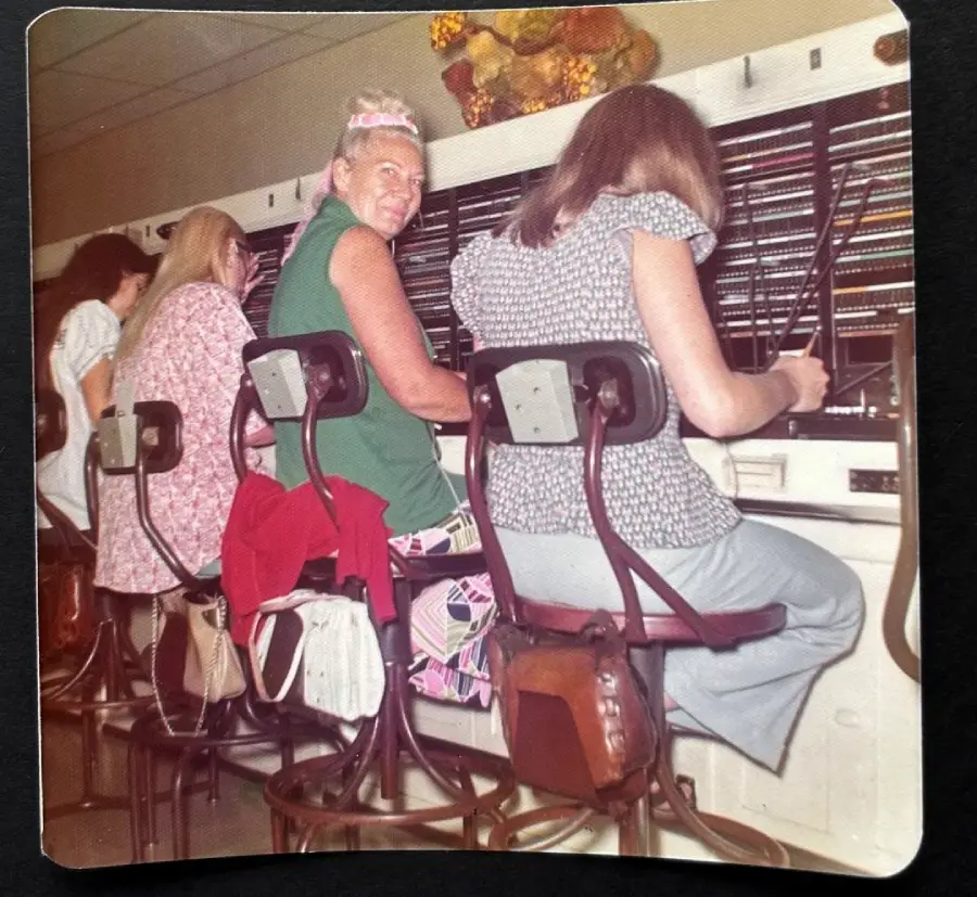 Four women are sitting on stools at a vintage telephone switchboard, working with cables and headsets. One woman in a green dress looks back and smiles at the camera. The scene appears to be from the 1970s.