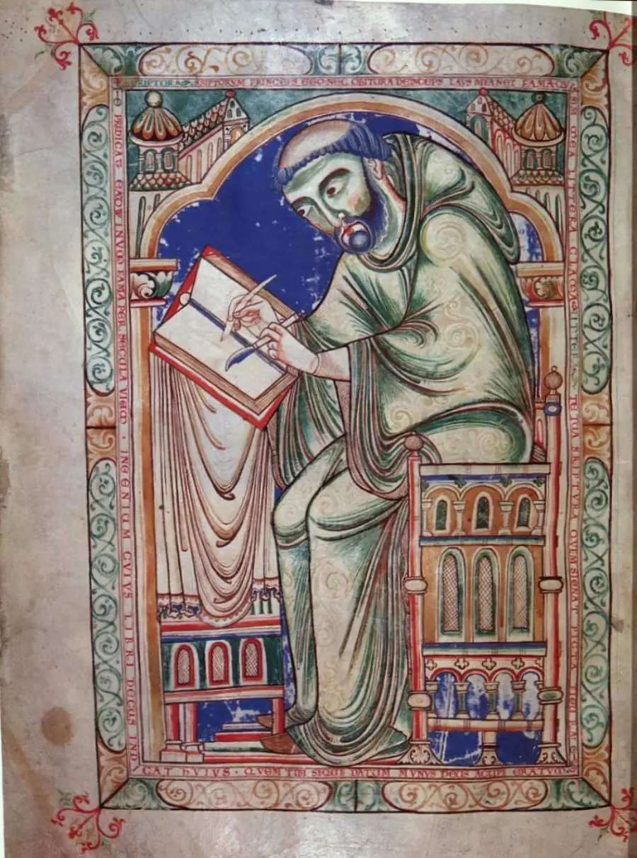 Medieval illustration of a robed monk seated at a desk, writing in a large book. The scene is framed with ornate arches and decorative borders in red, green, and blue.