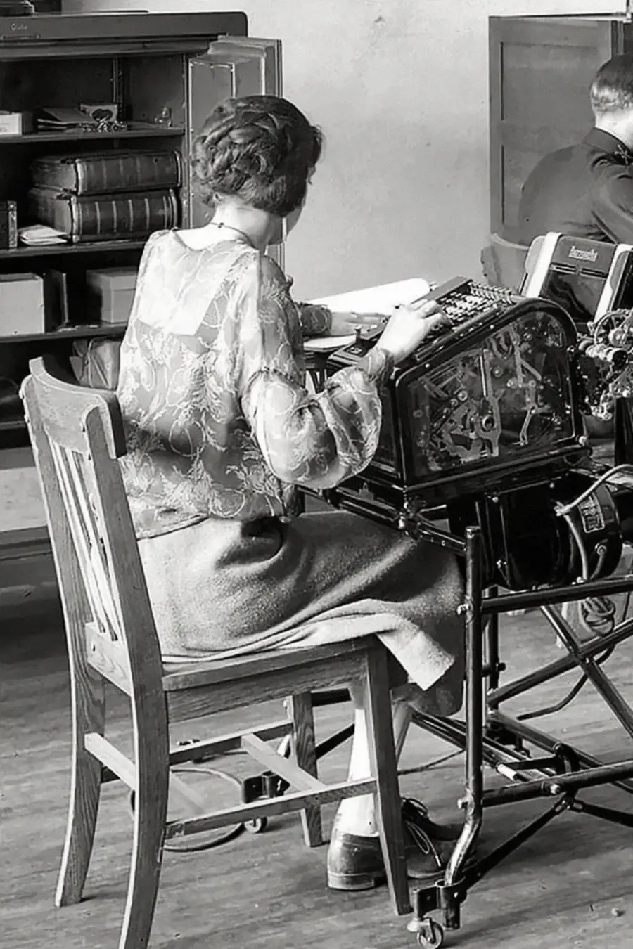 A woman sits at a wooden desk operating a vintage mechanical calculator, surrounded by paperwork. She wears a patterned blouse and skirt, with her hair neatly styled, in an office setting.