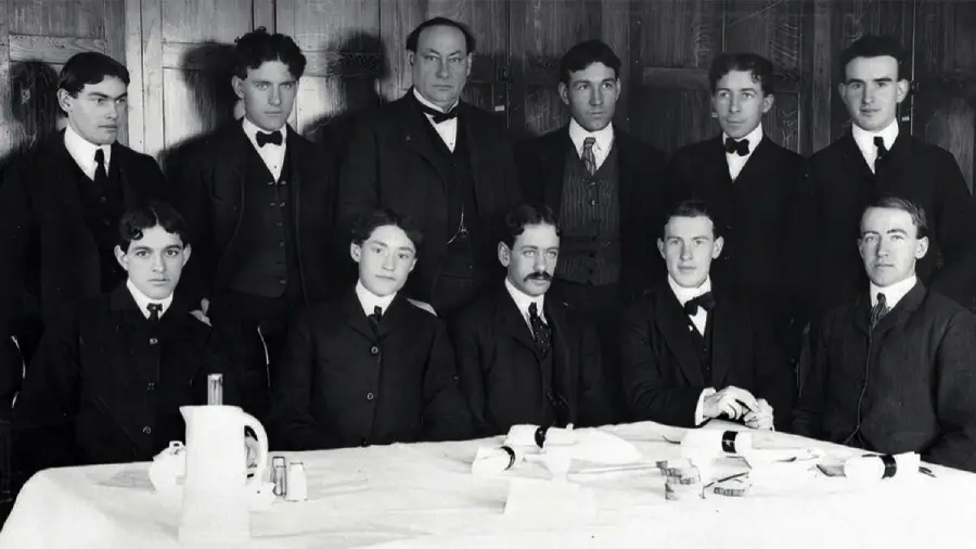 Black and white photo of eleven men in suits and ties, some seated at a table with a white cloth, papers, cups, and a pitcher, while others stand behind them, all posing for a formal group portrait.