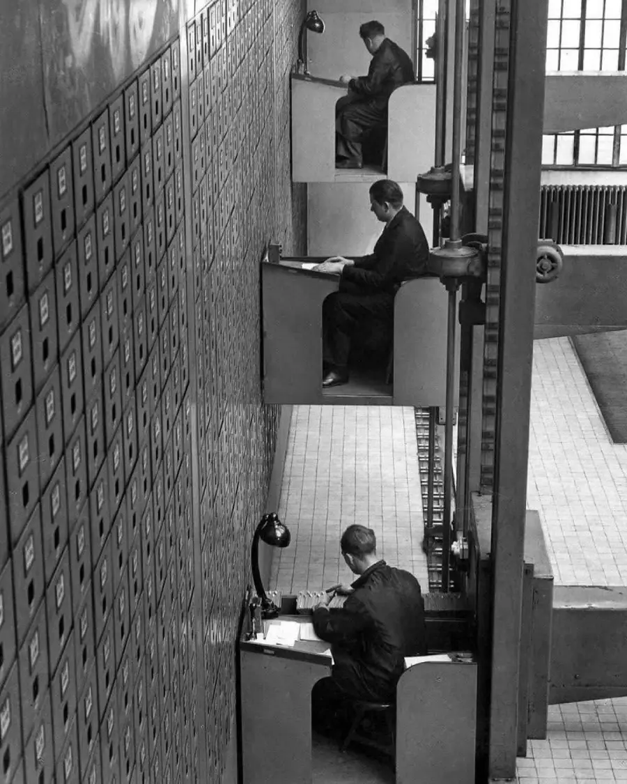 Three men sit at separate workstations built into a tall wall of drawers, each desk stacked vertically one above the other, working with papers and lamps in an industrial, office-like setting.