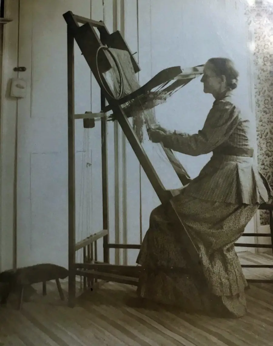 A woman in vintage clothing sits at a large upright loom, weaving. The setting appears old-fashioned, and a small animal, possibly a cat or dog, is on the floor nearby.