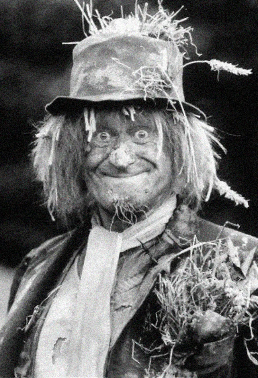 A person dressed as a scarecrow with straw in their hat and clothes, messy hair, a painted face, and a big grin, wearing a weathered suit and scarf. The image is in black and white.