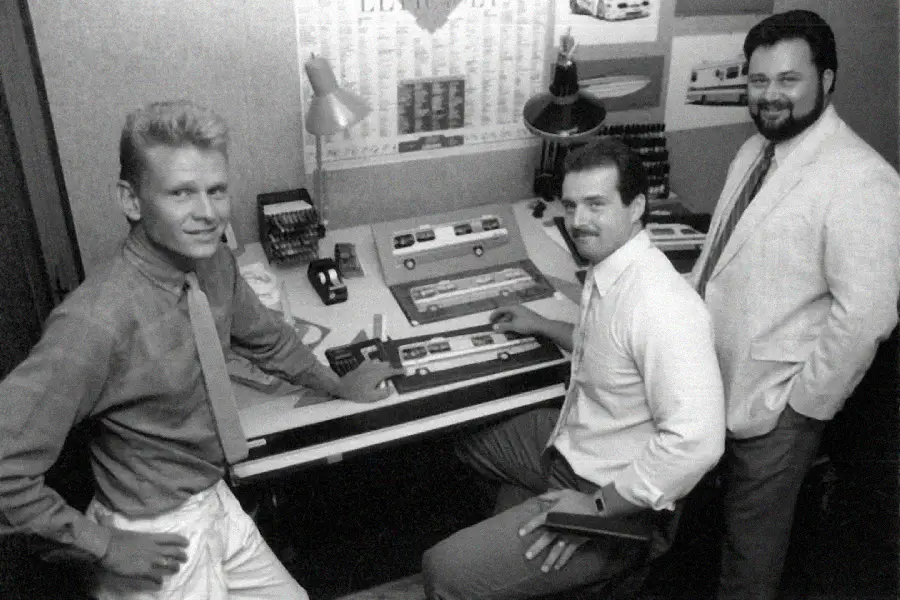 Three men in business attire pose around a drafting table with model vehicles and design tools. Behind them is a lamp, office supplies, and a poster featuring images of vehicles. They are smiling and looking at the camera.