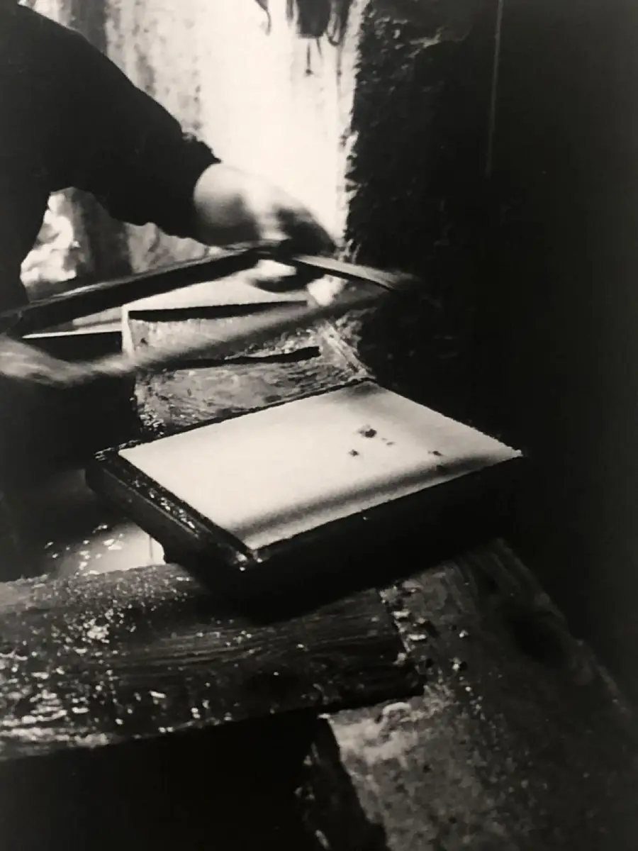 A person is making paper by hand, using a wooden frame to lift a thin layer of pulp from a vat, with water dripping onto a work surface.