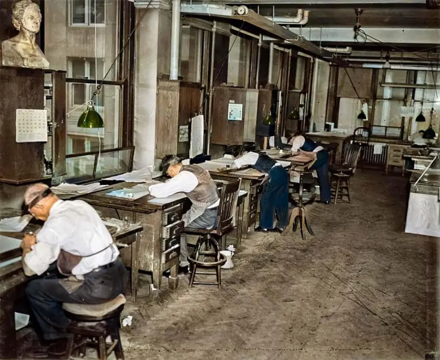 Several men wearing vests and visors work at drafting desks in an old-fashioned office with large windows, wooden partitions, and overhead lights. The room has a vintage, early 20th-century atmosphere.