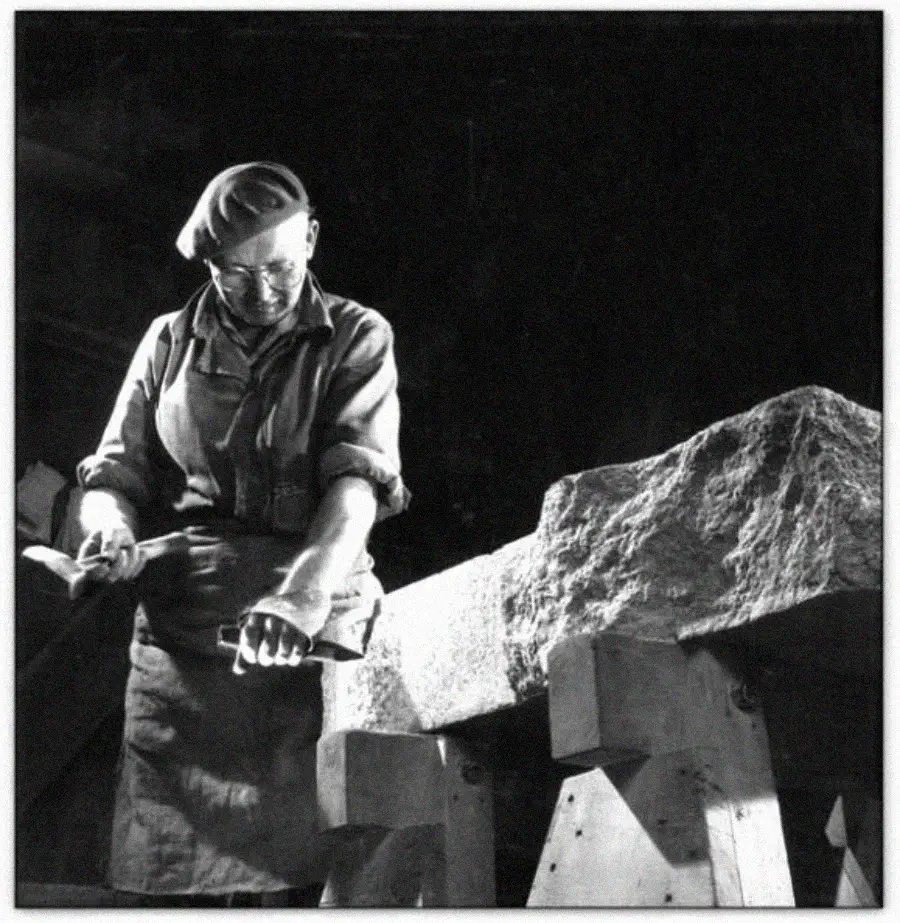 A person wearing glasses, a beret, and work clothes carves a large stone block with a hammer and chisel, standing in a workshop with dramatic lighting.