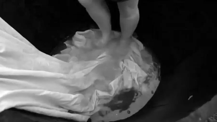 A person stands barefoot in a shallow container of water, pressing down on a wet sheet or cloth with their feet. The image is in black and white.
