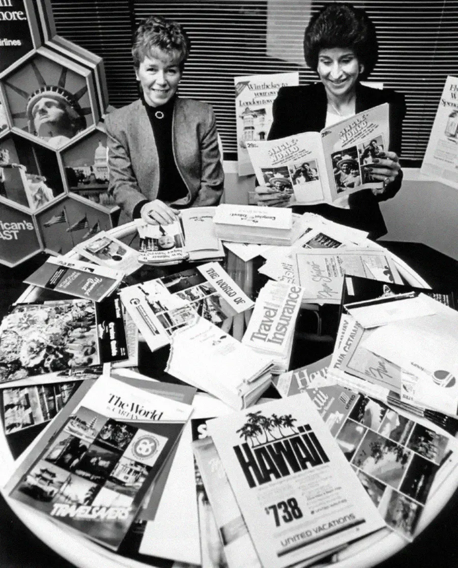 Two women sit at a round table covered with travel brochures and pamphlets, smiling as they look at vacation materials. Behind them is a display with more travel brochures and an image of the Statue of Liberty.
