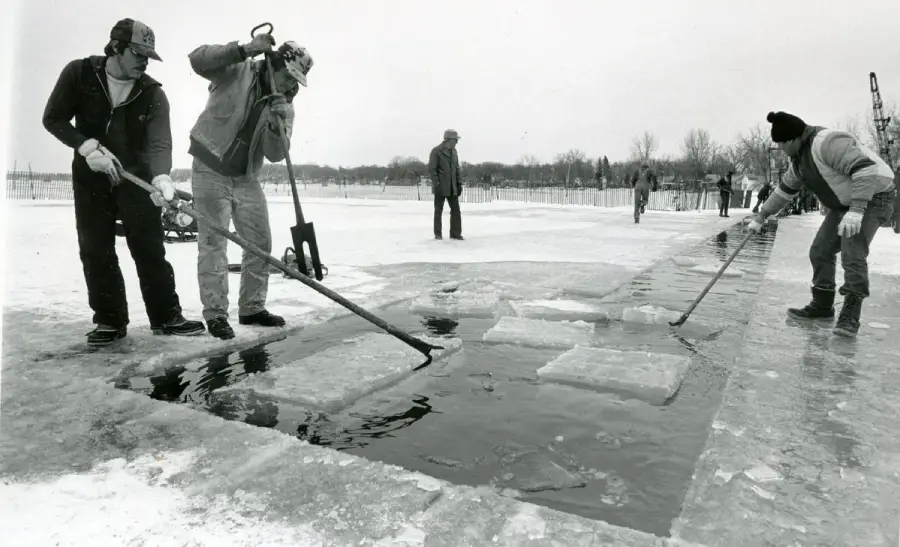 Three people in winter clothing use tools to lift and move blocks of ice from a frozen surface, while others stand in the background on a snowy landscape.