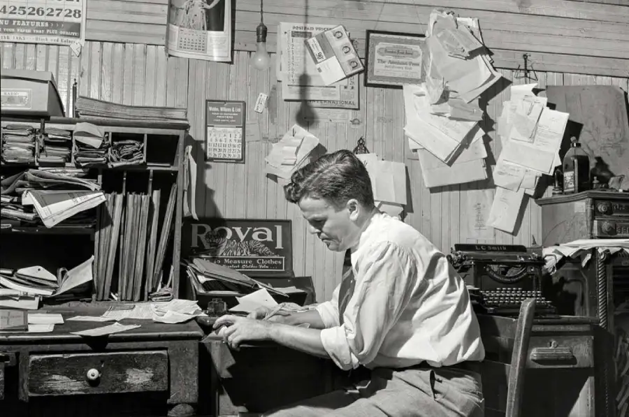 A man in a white shirt sits at a cluttered desk, surrounded by stacks of papers, a typewriter, and documents pinned to the wall in a busy, old-fashioned office.