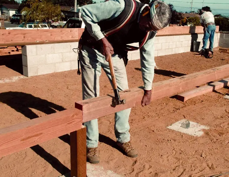 A person wearing work clothes and a headband uses a hammer to drive a nail into a wooden beam at a construction site with sandy ground and partially built walls.