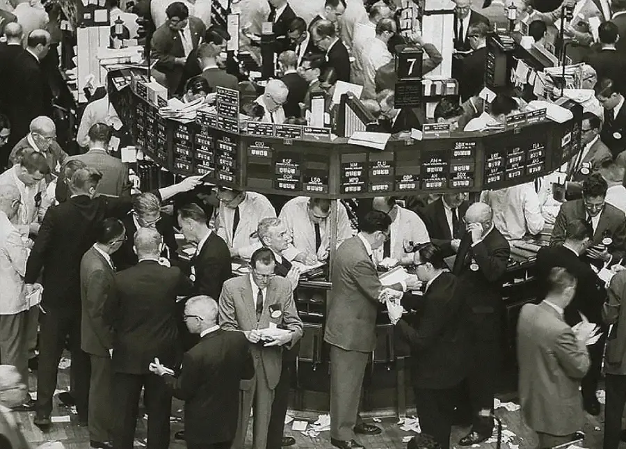 A crowded trading floor filled with men in suits, bustling around rows of trading booths and monitors, exchanging papers and transactions in a busy, hectic financial environment.