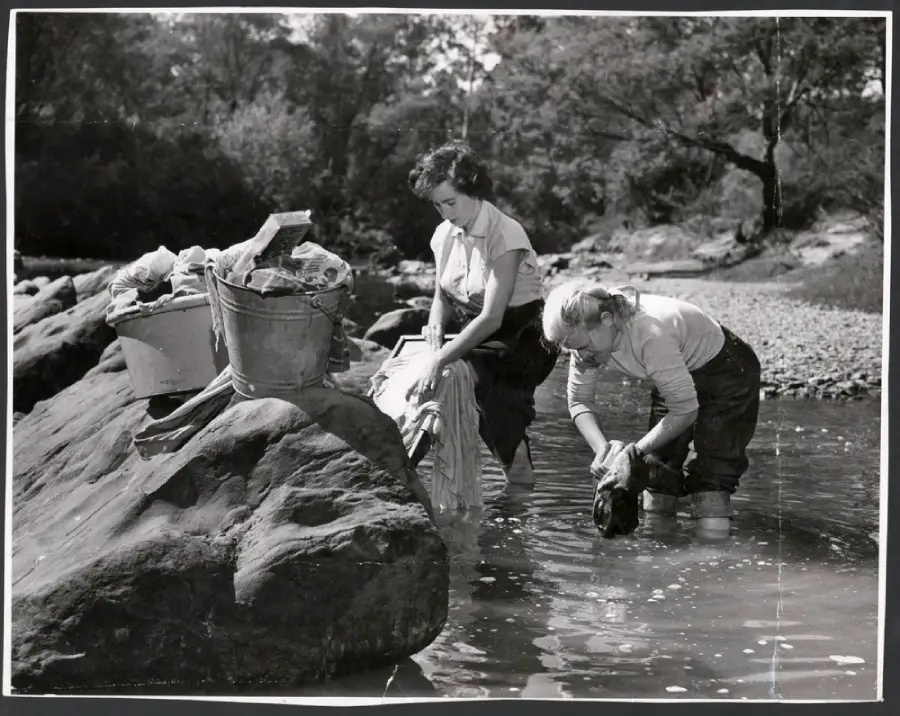 Two women wash clothes by hand in a shallow river. Laundry and buckets rest on a large rock nearby. Trees and rocks line the background under a clear sky.