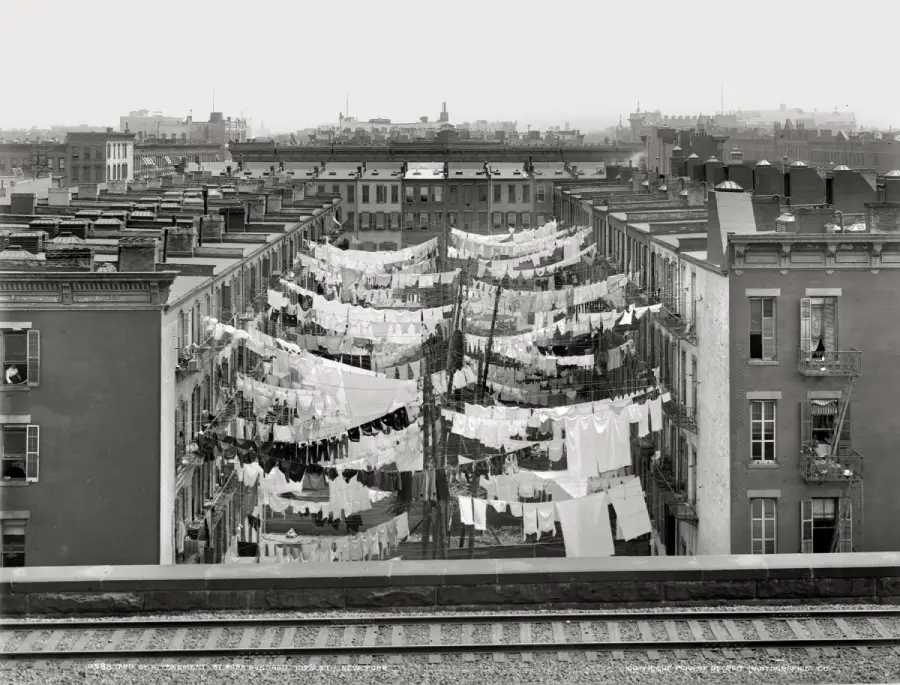 Rows of laundry hang on clotheslines strung between tenement buildings above a narrow alleyway in a dense city neighborhood, with rooftops and train tracks in the foreground.