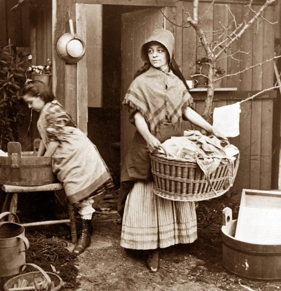 Two women do laundry outside. One washes clothes in a wooden basin, while the other, wearing a bonnet and shawl, carries a large basket of laundry. The scene appears to be from the late 19th or early 20th century.