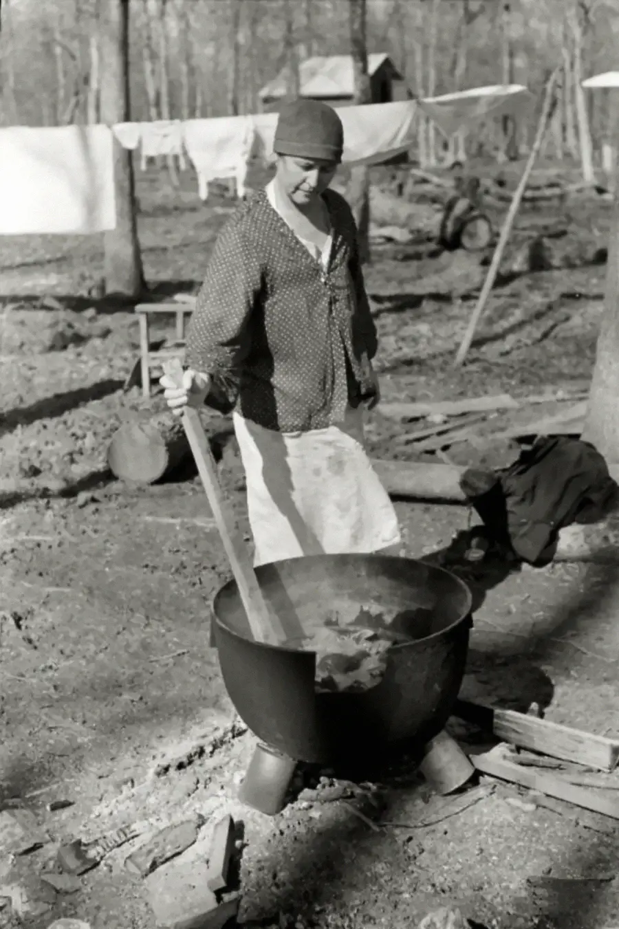 A person wearing a hat and polka dot shirt stirs a large pot outdoors. Laundry hangs on a line in the background, with scattered wood and trees around.
