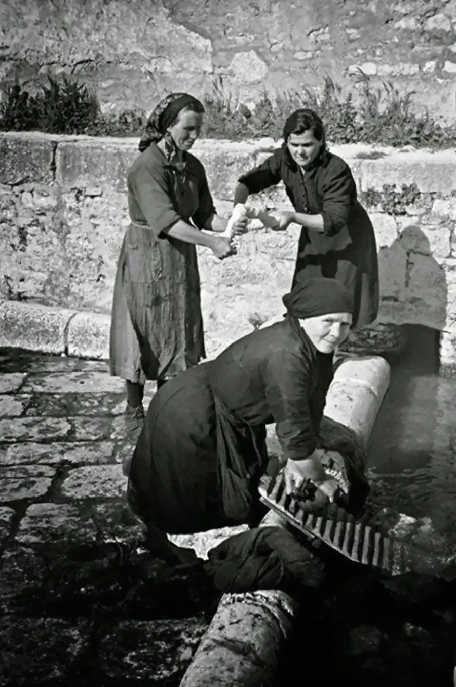 Three women in headscarves and dresses wash clothes by hand at an outdoor stone washbasin. One scrubs on a washboard while the other two twist laundry together. The setting appears historic or rural.