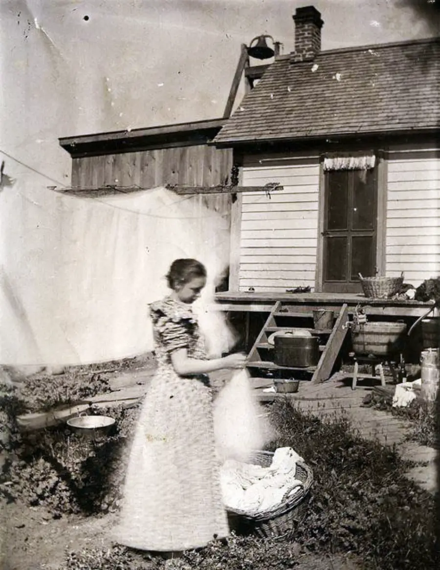 A woman in a patterned dress stands outside by a basket of laundry in front of a wooden house, with a wash line and various buckets in the yard. The photo is black and white, with a blurred motion effect.
