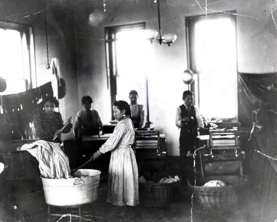 Women and children in an old-fashioned laundry room, washing and wringing clothes by hand. Large windows let in light. Several laundry baskets and tubs are visible. The photo appears worn and aged.