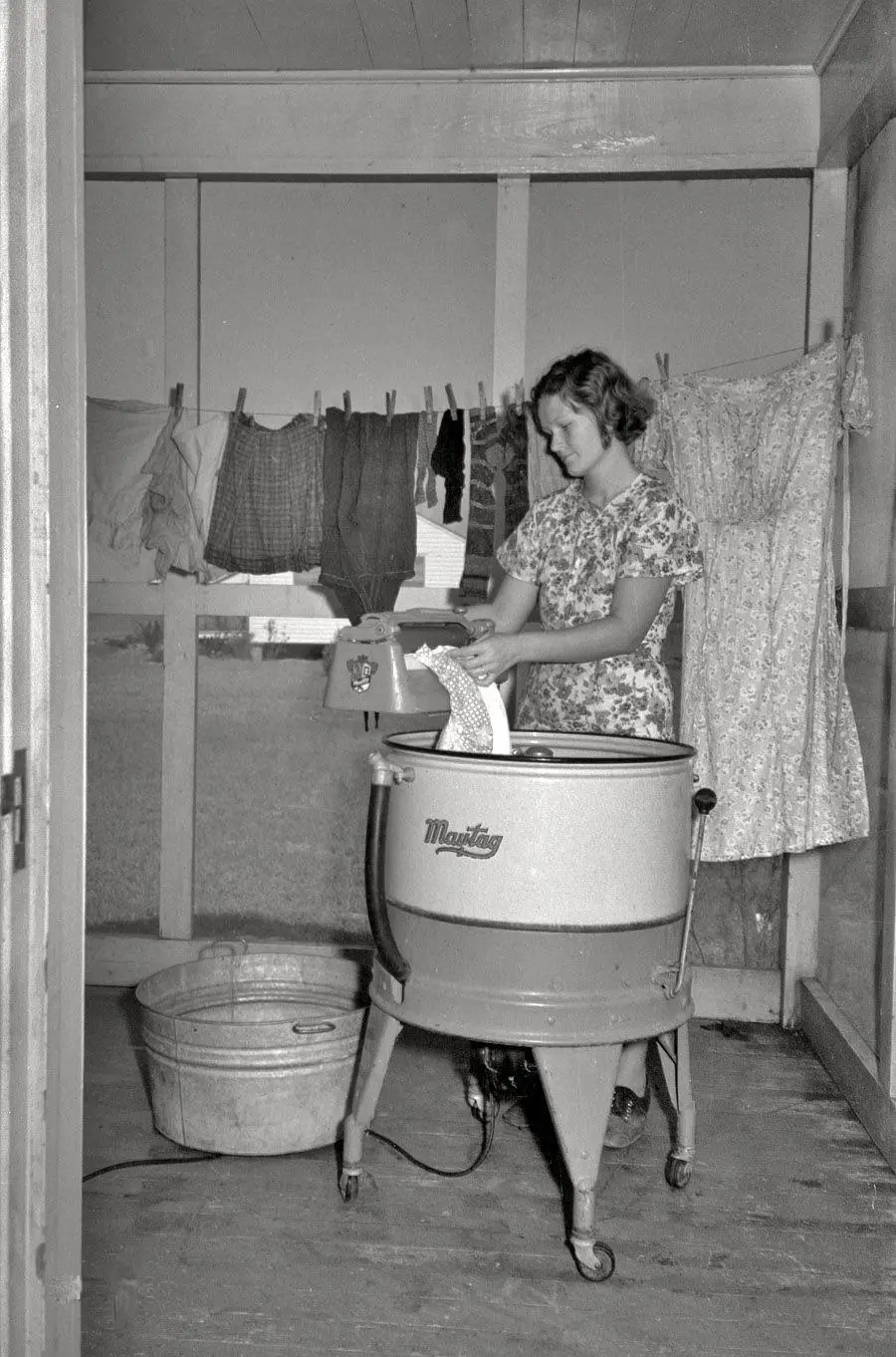 A woman stands by a vintage Maytag washing machine, wringing clothes. Laundry hangs on a line behind her, and a metal wash tub sits on the floor nearby. The scene is on an enclosed porch.