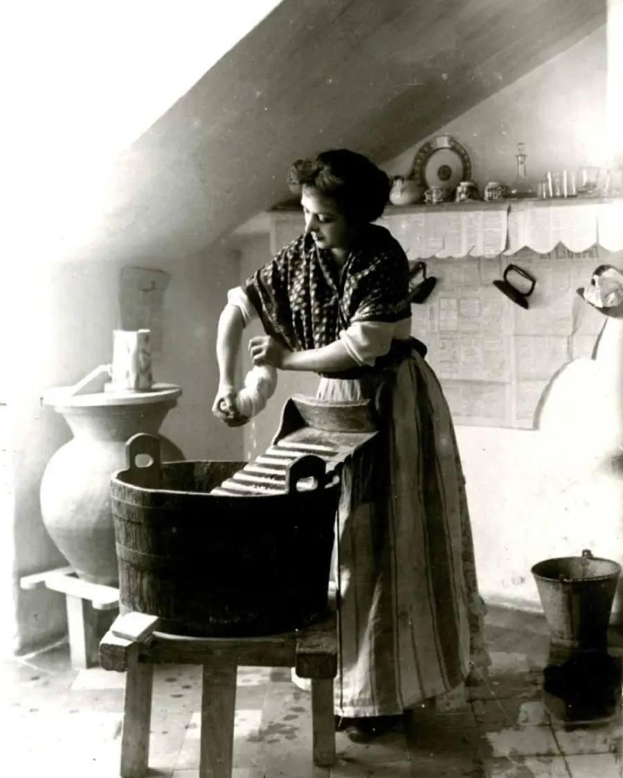 A woman in old-fashioned clothing washes clothes on a washboard in a wooden tub, set in a vintage kitchen with shelves, jars, and utensils in the background.