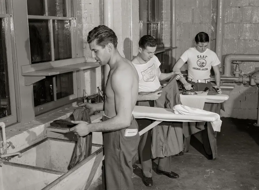 Three men iron clothes in a laundry room. One is shirtless and ironing at the front, while two others, wearing T-shirts, iron beside him. Sinks and pipes are visible in the background.