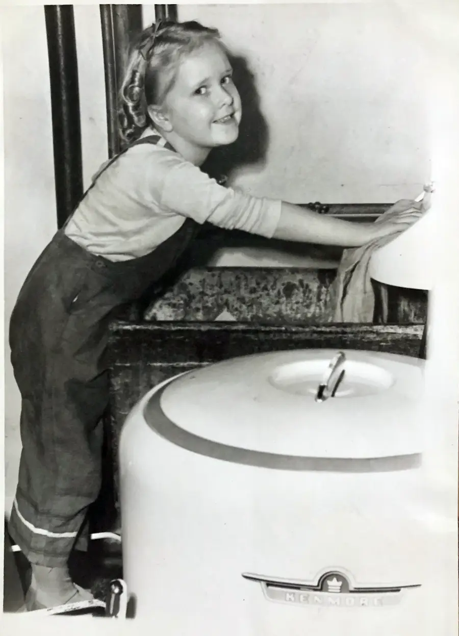 A smiling young girl in overalls washes clothes by hand in a sink next to a vintage Kenmore washing machine.