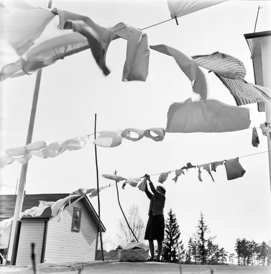 A person hangs laundry on a clothesline outdoors. Sheets and clothes billow in the wind, with a house and trees visible in the background under a bright sky.