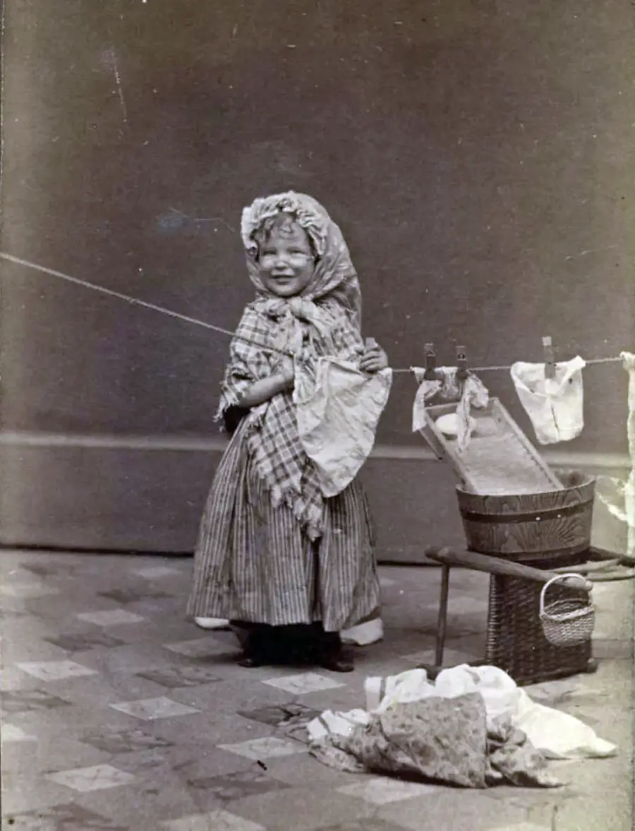 A young girl in old-fashioned clothes stands indoors, smiling. She hangs laundry on a line, with a basket, chair, and cloth pile nearby. The background is plain.