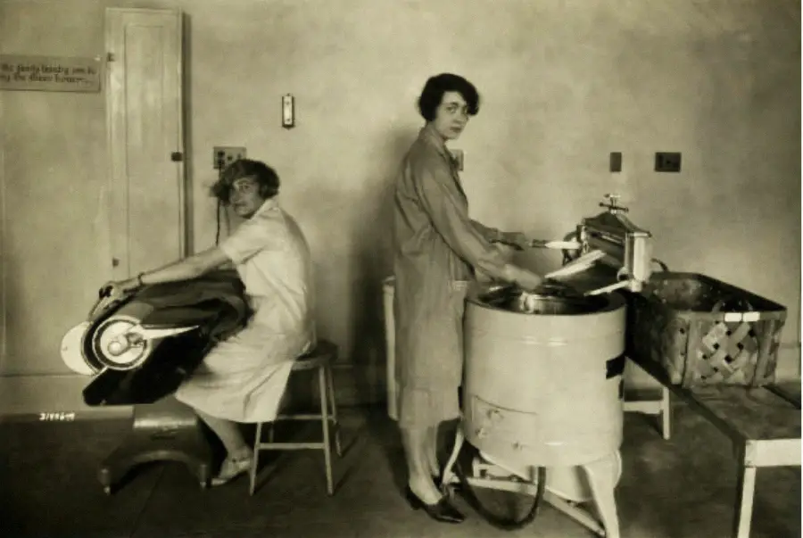 Two women in dresses use vintage laundry machines in a plain room—one operates a washing machine, while the other uses a wringer. Both women look toward the camera.