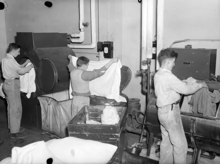 Three boys work in a laundry room, handling large sheets and placing them into industrial washing machines and bins. Pipes and equipment are visible in the background, suggesting a busy work environment.